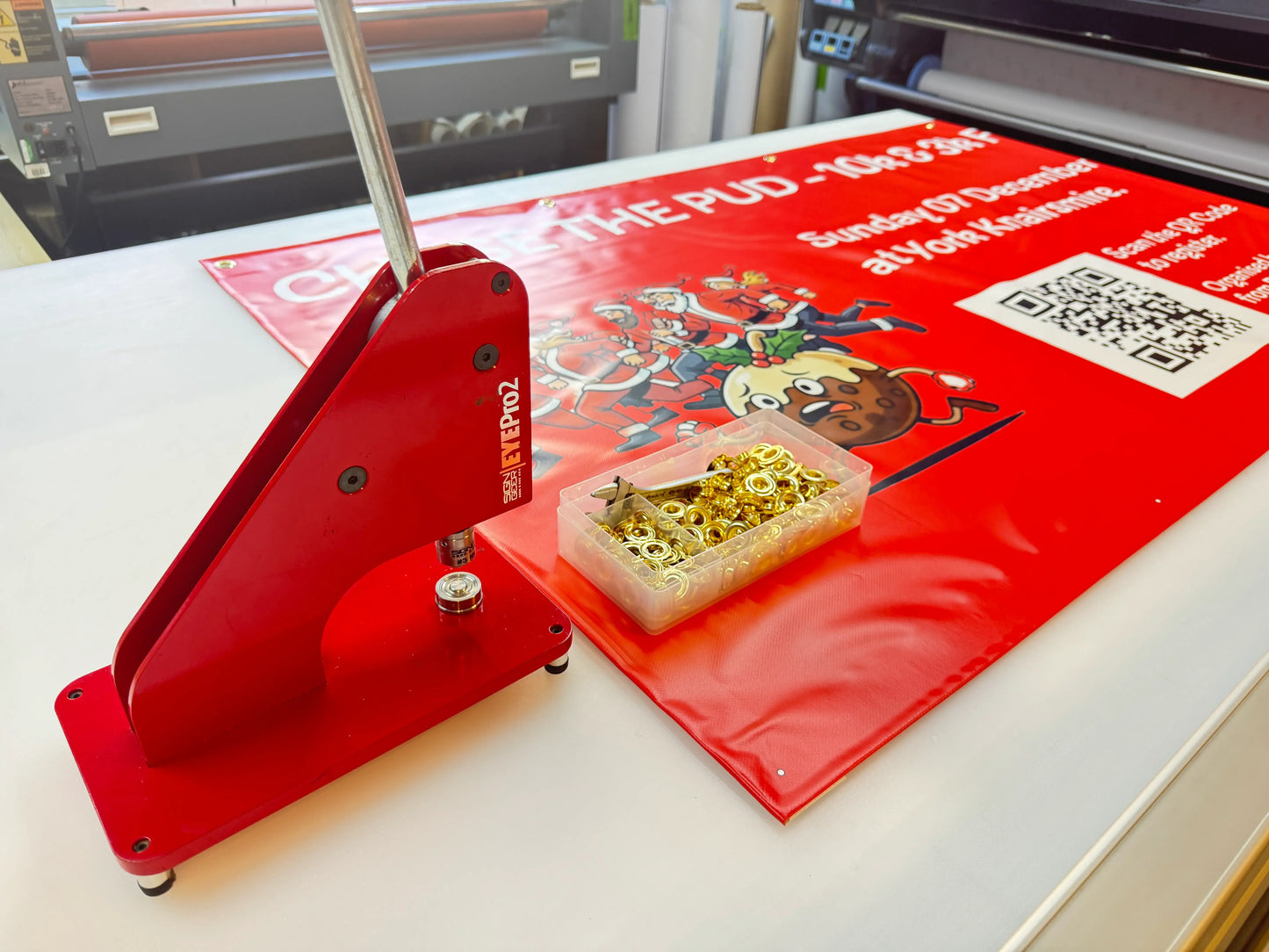 Red grommet press and eyelets beside a printed event banner on a print shop table